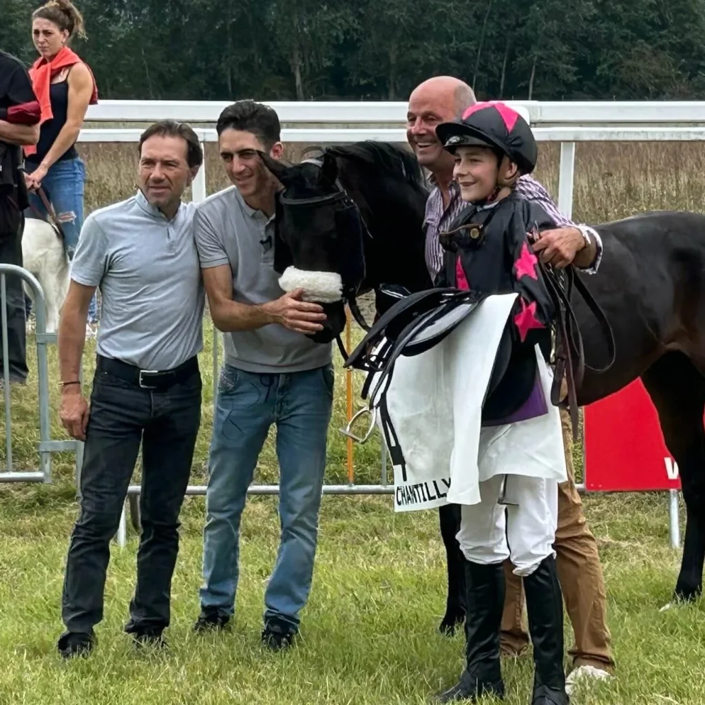 Jeune jockey souriant tenant sa selle à côté d'un cheval noir et de trois hommes à Chantilly