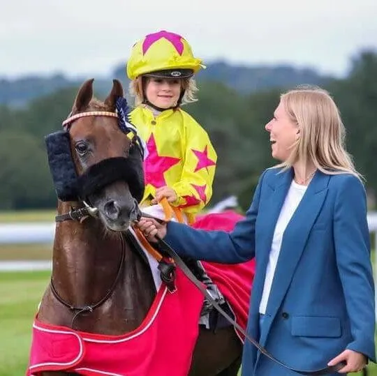Enfant en tenue de jockey colorée sur un poney, accompagnée d'une femme en veste bleue sur un hippodrome.
